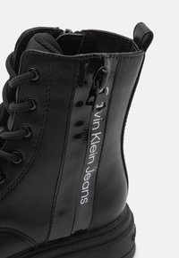 Black ankle boot made of smooth and patent leather, featuring textured fabric interior, lace-up front, and side zipper with logo detail.