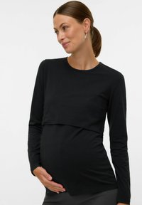 Trudnica s smeškom, smeškom nosi smešu smešu smešu smešu smešu smešu smešu smešu smešu smešu gori, noseći crnu majicu dugih rukava tečnog mečera. Drži rukom trbuh i blago se smeška.