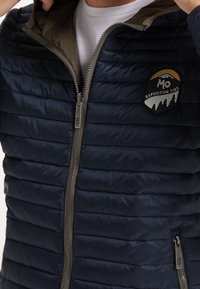 Veste matelassée marine avec des coutures horizontales, équipée d'une capuche contrastante vert olive. Soulignée par un patch et une fermeture zippée à l'avant.