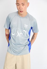 Světlá šedá sportovní dres s modrými akcenty, obsahující bílé nápisy "OBEY", vyrobený z prodyšného materiálu. Nosič má na sobě béžové kostkované šortky.