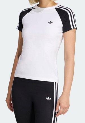 Kobieta nosząca biało-czarną koszulkę Adidas z trzema paskami na rękawach, w połączeniu z czarnymi legginsami z trzema białymi paskami i logo.