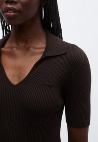 Polo marrón de punto acanalado con cuello en V y mangas cortas, con un pequeño logo bordado en el lado izquierdo del pecho. Textura suave.