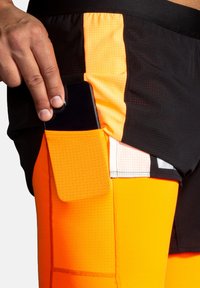Sportshorts i svart och klar orange, med en texturerad tyg. En orange ficka på sidan rymmer en smartphone.