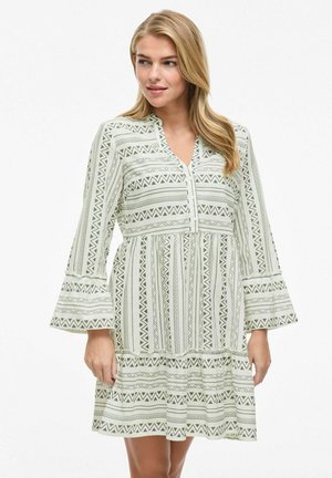 Robe verte à motifs avec un décolleté en V, des manches longues évasées et un ourlet à volants. Présente des motifs géométriques en noir et blanc.