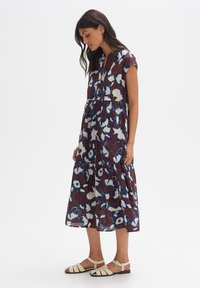 Blumenkleid in tiefem Burgunderrot mit blauen und weißen Mustern, ausgestattet mit einem gestuften Rock und kurzen Ärmeln. Kombiniert mit leichten Sandalen.