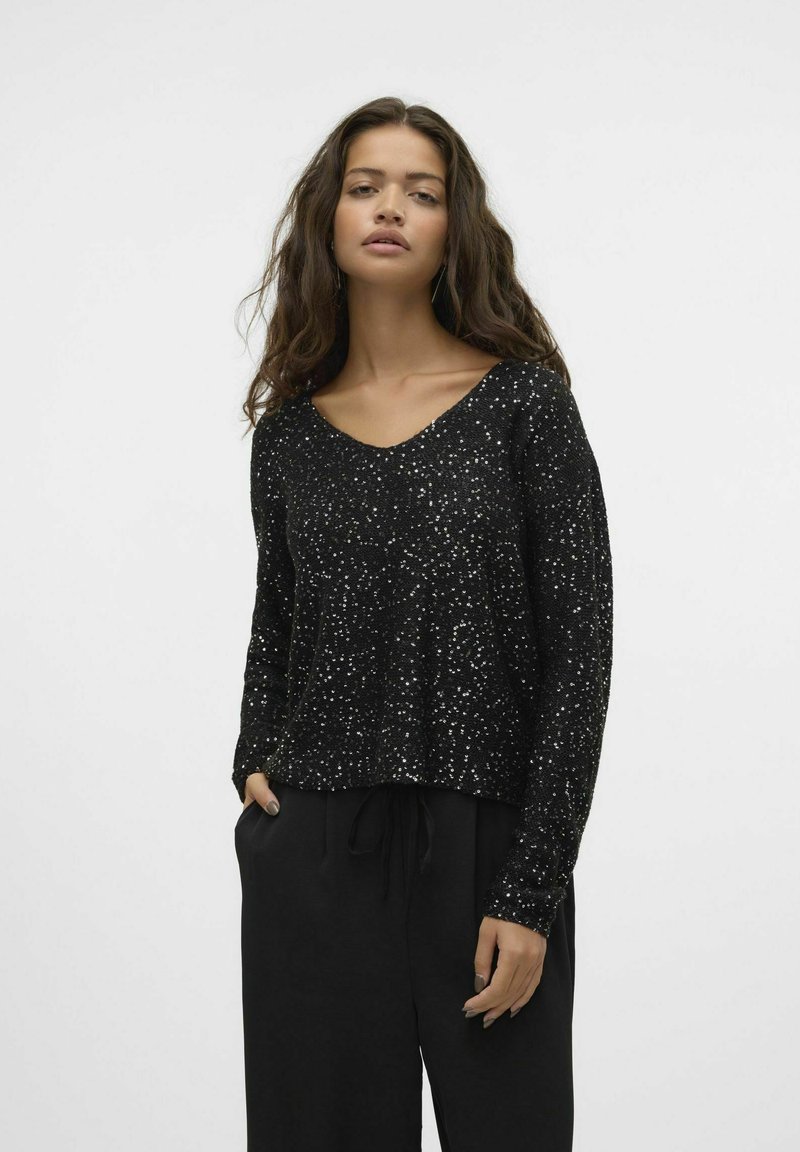 Maglione nero in paillettes con scollatura a V e maniche lunghe; vestibilità leggermente cropped. Indossato con pantaloni neri a vita alta per un look rilassato.