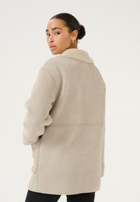 Beige tekokuitumainen suedesataviitta, jossa on karheapintainen vanhankermanvärinen shearling-kraavu, pitkät hihat ja rento istuvuus, jossa on puhtaat linjat ja vähäistä yksityiskohtaisuutta.