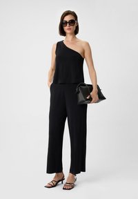 Schwarzes einseitiges Oberteil, kombiniert mit weit geschnittenen schwarzen Hosen. Hält eine kleine schwarze Handtasche. Trägt dazu schwarze Sandalen mit Absatz und Riemchen.