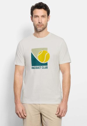 Uomo che indossa una t-shirt beige con pallina da tennis gialla e testo "Racquet Club", abbinata a pantaloni color sabbia chiaro, in piedi su sfondo bianco.