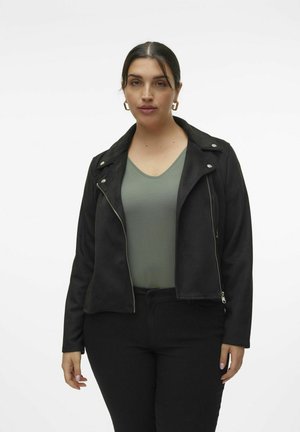 Veste en faux suède noir avec fermeture éclair et détails en argent, dotée d'un col cranté et d'une coupe ajustée. Portée par-dessus un top vert.