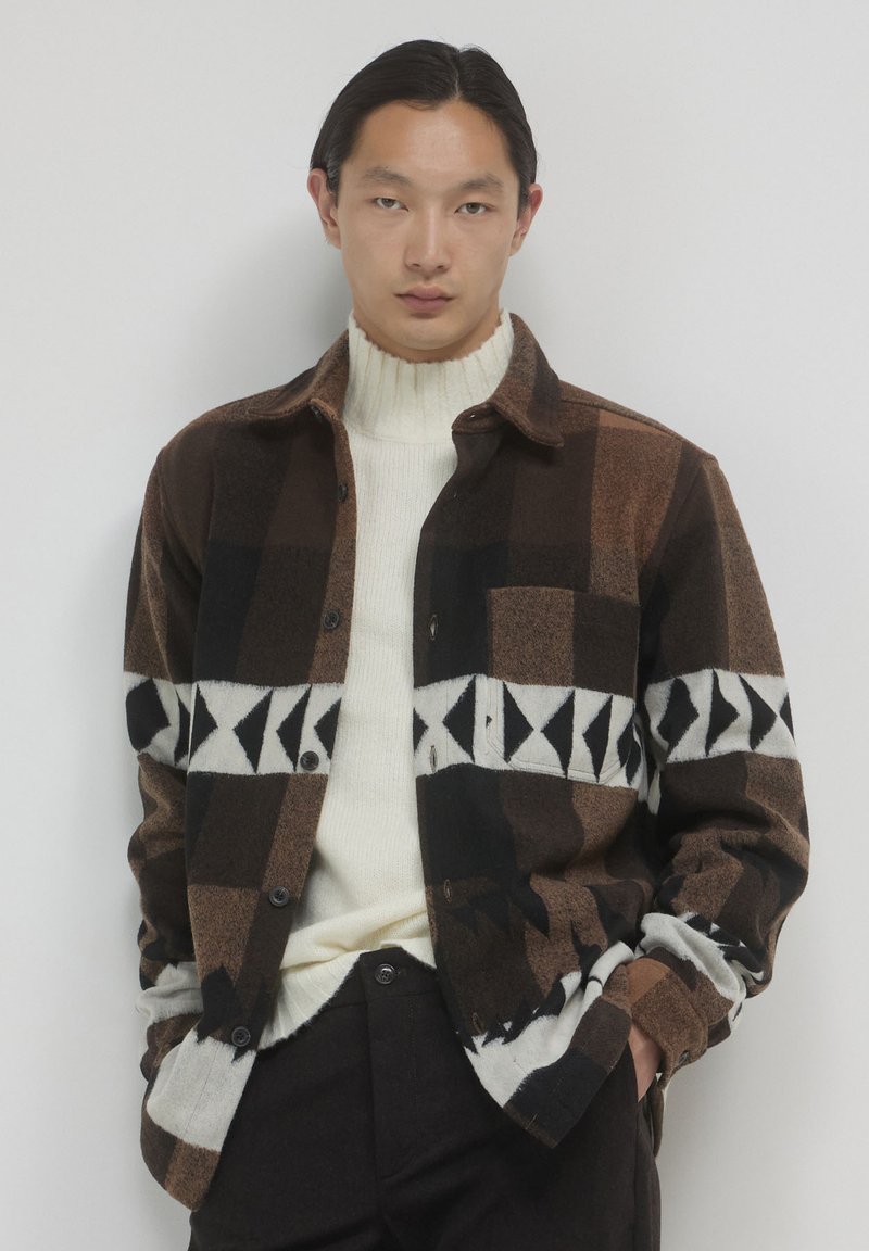 Chemise à carreaux brune et noire avec un motif géométrique blanc sur la poitrine, associée à un col roulé crème et un pantalon foncé.