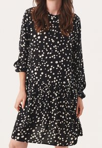 Abito nero con pois bianchi, maniche lunghe, pattino con bottoni e orlo a balze a strati. Realizzato in tessuto leggero.