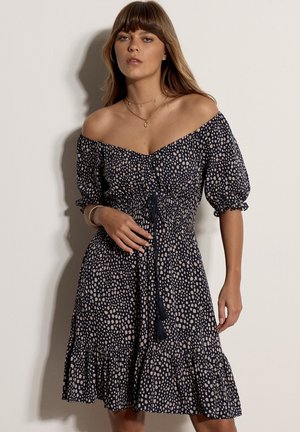 Kvinde iført off-shoulder navyblå kjole med beige pletter, elastisk talje med bindebånd, flæsekant og midt-lange puffede ærmer.