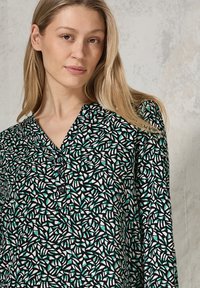 Blouse noire avec un motif géométrique de feuilles en vert et blanc, dotée d'un décolleté en V et de boutons à l'avant, fabriquée en tissu léger.
