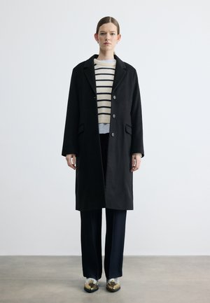 Woman stands wearing a long black coat, cream and navy striped sweater, black pants, and metallic gold shoes on plain background.