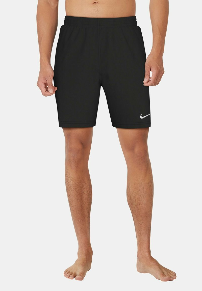 Άνδρας που φοράει μαύρα αθλητικά σορτς Nike, όρθιος ξυπόλητος μπροστά σε απλό φωτεινό φόντο.