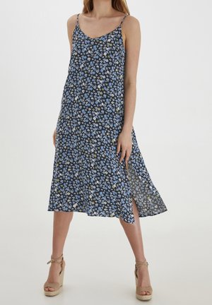 Vrouw draagt een zwarte midijurk met blauw-witte bloemenprint, spaghettibandjes en beige sleehakken met enkelbandjes.