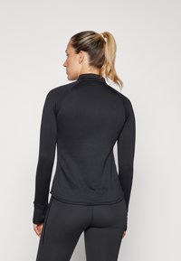 Fekete, hosszú ujjú sportfelső magas gallérral és bordázott textúrával, fekete, testhezálló leggingsszel párosítva, hátulról nézve.