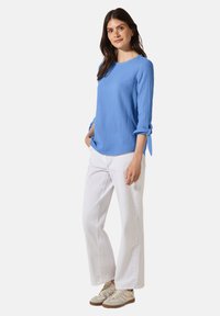 Maglietta azzurro chiaro a maniche lunghe con polsini annodati, abbinata a pantaloni bianchi a gamba larga e sneakers beige, indossata da un modello su uno sfondo semplice.