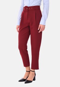 Pantaloni bordeaux a vita alta con una cintura larga dotata di una fibbia rettangolare, pieghe strutturate e caviglie affusolate, abbinati a tacchi neri.