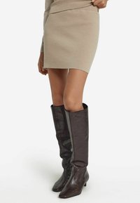 Kniehoge bruine leren laarzen met een glanzende textuur, een vierkante neus en een lage hak, gecombineerd met een aansluitende beige gebreide rok.