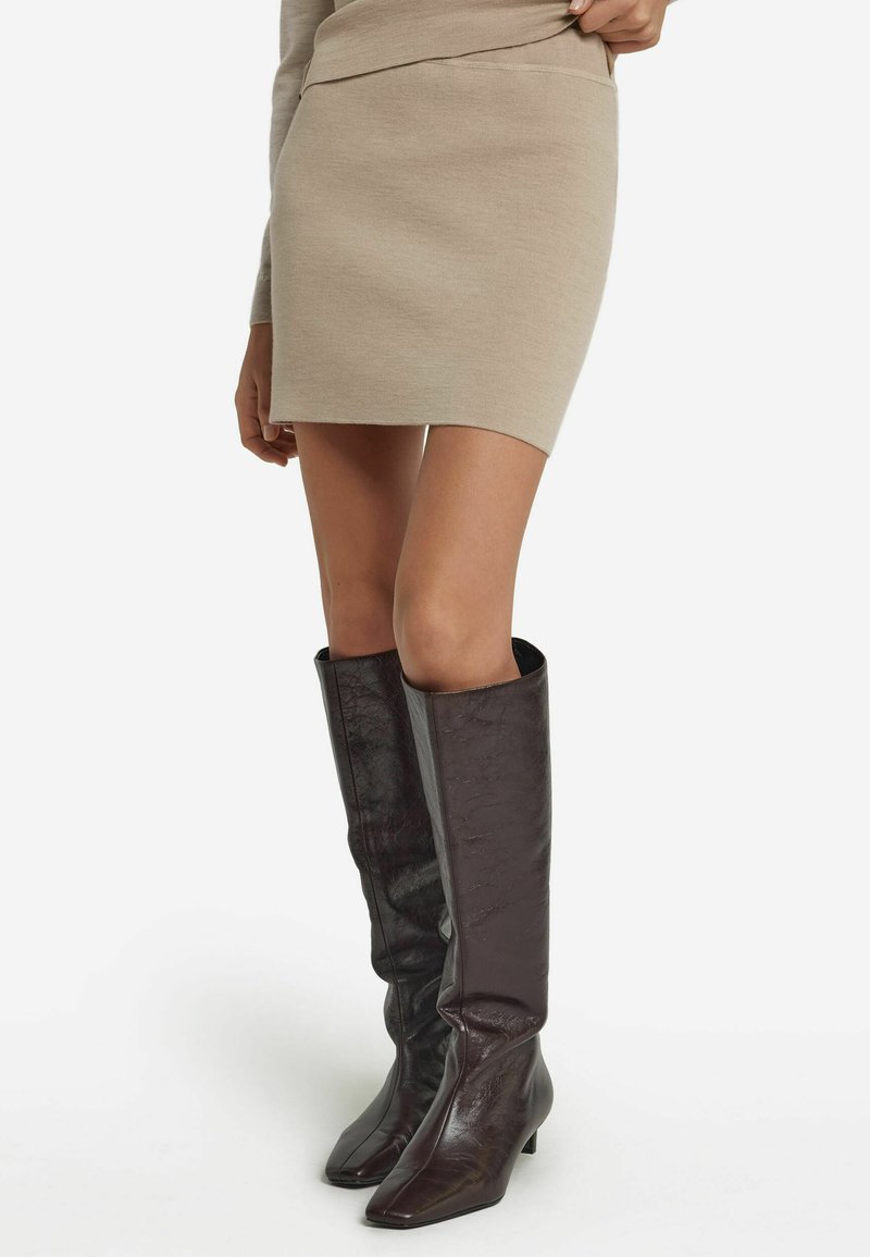 Kniehoge bruine leren laarzen met een glanzende textuur, een vierkante neus en een lage hak, gecombineerd met een aansluitende beige gebreide rok.