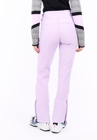 Lazdų spalvos slidinėjimo kelnės su lieknu siluetu, turi šoninius užtrauktukus prie apačios ir glotnią, tamprią tekstūrą. Derinamos su juodai baltais raštuotais ilgomis rankovėmis.