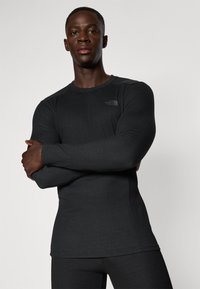 Juoda ilgomis rankovėmis atletinė marškinė su prigludusia forma, lygia tekstūra ir subtiliu prekės ženklu ant krūtinės, demonstruojama su sukryžiuotomis rankomis.