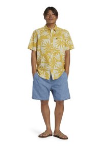 Camicia gialla chiara a maniche corte con motivo a foglie bianche, con bottoni davanti, abbinata a pantaloncini di jeans e ciabatte. Stile casual da spiaggia.
