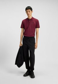 Maroon Polohemd mit schwarzem Besatz, drei Knöpfen und Logo. Schwarze Jogginghose mit Kordelzug; das Modell hält eine schwarze Jacke. Schwarze Sneaker.