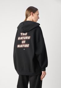 Svart hettegenser med oversized passform, med teksten "THE NATURE OF NATURE" i slitt hvitt. Mykt materiale med kengurulomme og snøring på hetta.