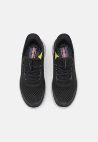 Czarne sportowe buty typu slip-on z przewiewną siatkową cholewką, teksturowanym wykończeniem i szarym podbiciem. Żółty akcent przy pięcie.