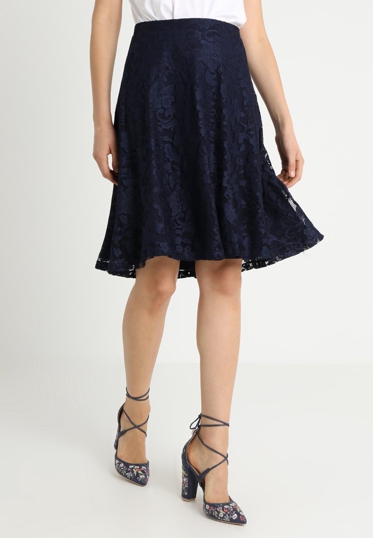 Femme portant une jupe trapèze en dentelle florale bleu marine et des chaussures à talons hauts bleu marine avec broderies florales et brides à la cheville.