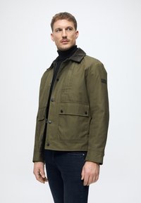 Jaqueta verde-azeitona feita de lona, com uma gola de veludo cotelê preta, bolsos frontais e fechos de botão. Usada sobre um suéter de gola alta preto.