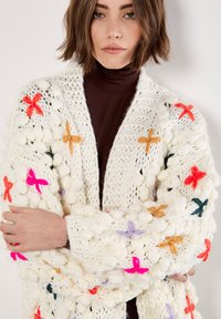 Cardigan bianco lavorato a maglia con farfalle ricamate colorate in arancione, rosa e verde. Presenta una vestibilità ampia e oversize e un motivo testurizzato.