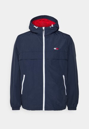 Granatowa kurtka z kapturem z czerwoną podszewką, białym zamkiem błyskawicznym, elastycznymi mankietami, dwoma zamykanymi na zamek bocznymi kieszeniami i logo Tommy Jeans na piersi.