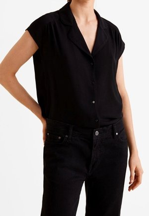 Blouse noire à manches courtes avec col, fermeture à boutons à l'avant et texture de tissu lisse, associée à un jean noir avec poches classiques et boutons.