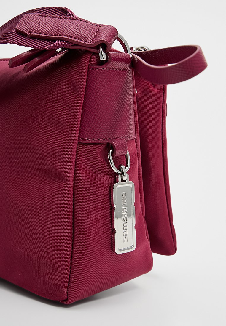Bolsa de nailon color marrón con un acabado texturizado, que presenta una etiqueta plateada y una correa. El diseño incluye un cierre en la parte superior y una forma angular.