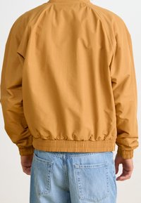 Bombarjacka i senapsgult, tillverkad av mjukt tyg. Har ribbat nederkant och ärmmuddar med en avslappnad passform. Bärs över ljusblå jeans.