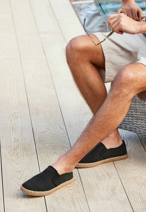 Crne slip-on cipele s teksturiranom gornjom stranom i tkanim jute potplatom, smještene na svijetlom drvenom podu. Nema vidljivih brandova ili uzoraka.