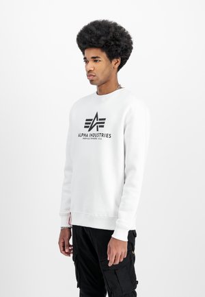 Lys grå sweatshirt med sort "Alpha Industries" logo, rund halsudskæring, ribbede manchetter og kant. Parret med sorte cargobukser.