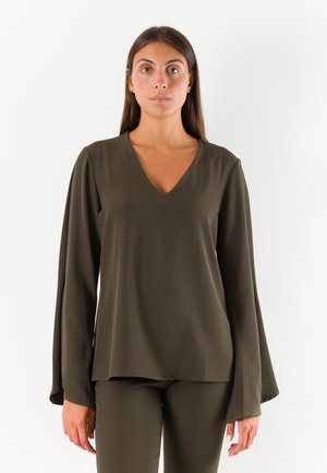 Donna con lunghi capelli castani che indossa una maglia verde oliva a maniche lunghe con scollo a V e pantaloni abbinati, in piedi davanti a uno sfondo bianco semplice.