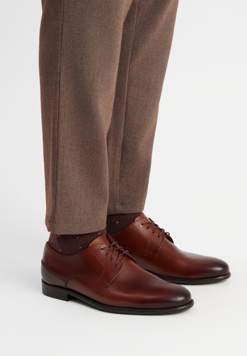 Zapatos de vestir de cuero marrón con un acabado suave, cordones y una forma elegante. Combinados con pantalones marrones texturizados y calcetines oscuros de color marrón con patrones.