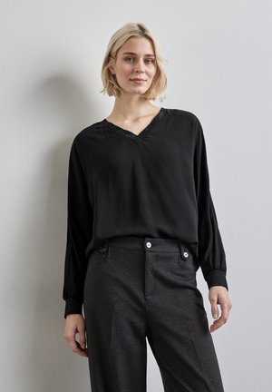 Svart långärmad blus med V-ringning och texturerad svart kant, tillsammans med grå mönstrade byxor med två knappar och rak skärning.