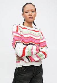 Νεαρό άτομο με πλεξούδες, που φοράει πολύχρωμο ριγέ πλεκτό πουλόβερ και σκούρο τζιν, με σταυρωμένα τα χέρια, στέκεται μπροστά σε ένα απλό φωτεινό φόντο.