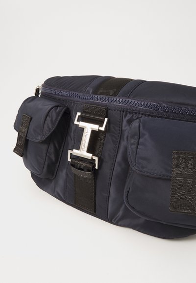 Navy blue nylon bag with a smooth texture, featuring two front pockets, a silver buckle, and a black strap. Zip closure at the top.
