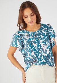 Blusa de manga corta con un patrón floral en verde azulado sobre un fondo blanco, con detalles de botones y un corte relajado. Textura de tela suave.