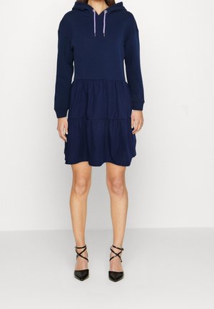 Femme portant une robe à capuche bleu marine à manches longues et jupe à volants, associée à des escarpins noirs à bout pointu avec brides à la cheville.