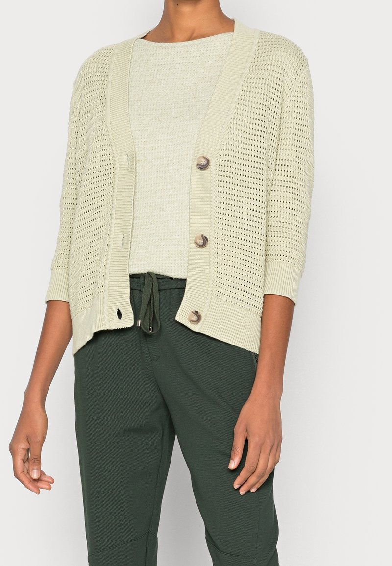 Femme portant un cardigan beige en maille ouverte boutonné sur un haut beige clair avec un pantalon vert foncé à cordon, devant un fond uni.