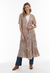 Robe longue à motifs paisley en tons beiges, roses et violets, avec des manches courtes, un décolleté en v et un ourlet en couches. Portée sur un jean et des bottes.